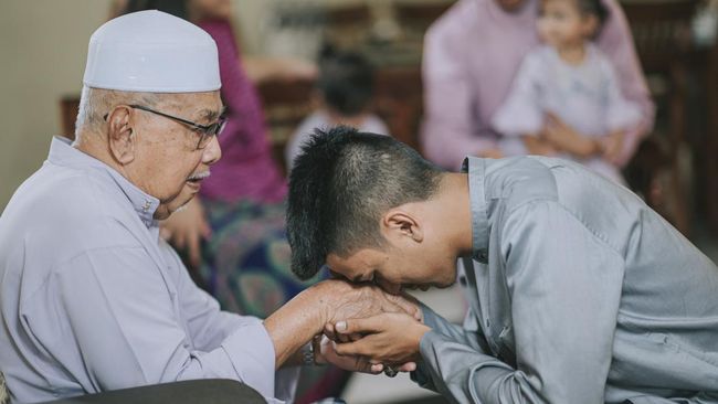 Seorang pemuda berbaju koko abu-abu sedang membungkuk mencium tangan seorang pria lansia berpeci putih dengan khidmat dalam suasana kekeluargaan.