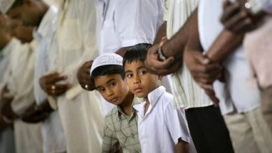 Dua anak laki-laki Muslim menatap ke kamera sambil berdiri di antara pria dewasa yang sedang shalat berjamaah di masjid.