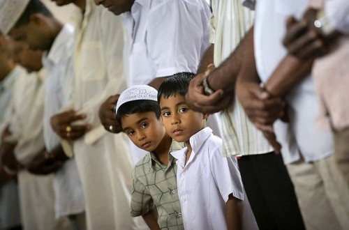 Dua anak laki-laki Muslim menatap ke kamera sambil berdiri di antara pria dewasa yang sedang shalat berjamaah di masjid.