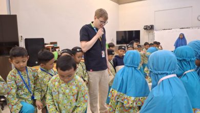 Seorang pengajar native speaker pria dengan kacamata sedang duduk bersama sekelompok siswa SD berseragam batik hijau dan jilbab biru, bersama-sama membaca dan mendalami buku teks "Pearson Global English Primary" yang dipegang oleh sang pengajar.