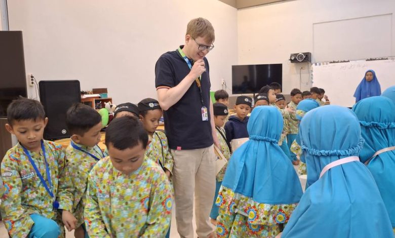 Seorang pengajar native speaker pria dengan kacamata sedang duduk bersama sekelompok siswa SD berseragam batik hijau dan jilbab biru, bersama-sama membaca dan mendalami buku teks "Pearson Global English Primary" yang dipegang oleh sang pengajar.