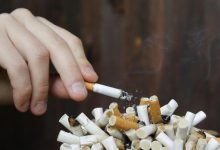 Tangan seseorang sedang memegang rokok yang menyala di atas tumpukan puntung rokok di dalam asbak yang penuh, menggambarkan sifat adiktif dan bahaya merokok.