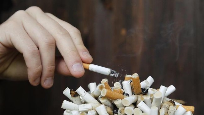 Tangan seseorang sedang memegang rokok yang menyala di atas tumpukan puntung rokok di dalam asbak yang penuh, menggambarkan sifat adiktif dan bahaya merokok.