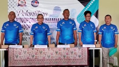 Foto lima pria berdiri di belakang meja panjang dalam seragam olahraga biru cerah. Di latar belakang terdapat spanduk bertuliskan "PELANTIKAN PINGPONG MUHAMMADIYAH KOTA PEKALONGAN PERIODE 2026-2030". Di atas meja terdapat tanda kertas bertuliskan (dari kiri ke kanan): "NARASUMBER 1", "DPRD JATENG", "NARASUMBER 2", "MODERATOR", dan "PENGURUS HARI". Pria di paling kanan memegang sebuah map atau dokumen cyan. Meja ditutupi taplak batik.