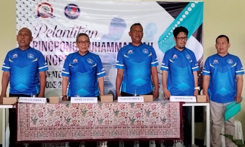 Foto lima pria berdiri di belakang meja panjang dalam seragam olahraga biru cerah. Di latar belakang terdapat spanduk bertuliskan "PELANTIKAN PINGPONG MUHAMMADIYAH KOTA PEKALONGAN PERIODE 2026-2030". Di atas meja terdapat tanda kertas bertuliskan (dari kiri ke kanan): "NARASUMBER 1", "DPRD JATENG", "NARASUMBER 2", "MODERATOR", dan "PENGURUS HARI". Pria di paling kanan memegang sebuah map atau dokumen cyan. Meja ditutupi taplak batik.