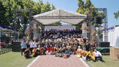 Foto bersama puluhan mahasiswa, panitia berkaus hitam, anggota pramuka, dan resimen mahasiswa di depan panggung acara outdoor.
