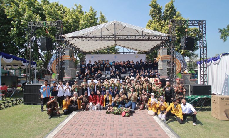 Foto bersama puluhan mahasiswa, panitia berkaus hitam, anggota pramuka, dan resimen mahasiswa di depan panggung acara outdoor.
