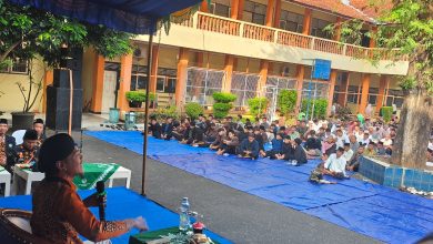 Seorang pembicara pria mengenakan batik oranye dan peci sedang berbicara di depan mikrofon di depan audiens besar yang duduk di lantai di atas terpal biru di halaman sekolah, dengan gedung sekolah oranye-kuning di latar belakang.