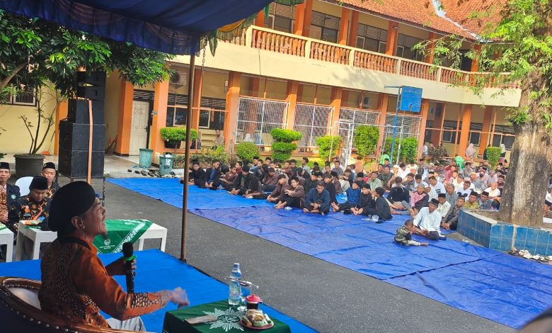 Seorang pembicara pria mengenakan batik oranye dan peci sedang berbicara di depan mikrofon di depan audiens besar yang duduk di lantai di atas terpal biru di halaman sekolah, dengan gedung sekolah oranye-kuning di latar belakang.