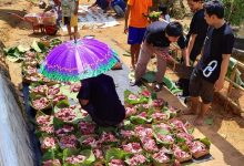 Grid keranjang anyaman hijau berisi daging qurban merah mentah yang tertata rapi di atas terpal, di bawah pengawasan sukarelawan dengan payung ungu di lokasi luar ruangan.