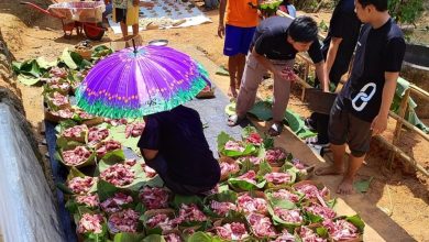 Grid keranjang anyaman hijau berisi daging qurban merah mentah yang tertata rapi di atas terpal, di bawah pengawasan sukarelawan dengan payung ungu di lokasi luar ruangan.