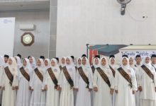 Sekelompok besar siswa-siswi SMP Muhammadiyah 2 Cilacap mengenakan seragam putih bersih, jilbab, songkok, dan slempang 'Wisuda Tahfidz' hitam, sedang melantunkan ayat suci Al-Qur'an secara koor di atas panggung di sebuah aula masjid. Mereka terlihat khidmat dan penuh konsentrasi saat diwisuda.