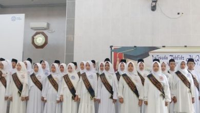 Sekelompok besar siswa-siswi SMP Muhammadiyah 2 Cilacap mengenakan seragam putih bersih, jilbab, songkok, dan slempang 'Wisuda Tahfidz' hitam, sedang melantunkan ayat suci Al-Qur'an secara koor di atas panggung di sebuah aula masjid. Mereka terlihat khidmat dan penuh konsentrasi saat diwisuda.