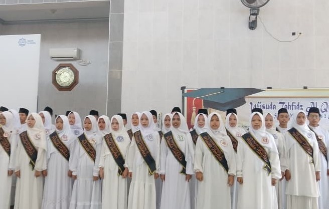Sekelompok besar siswa-siswi SMP Muhammadiyah 2 Cilacap mengenakan seragam putih bersih, jilbab, songkok, dan slempang 'Wisuda Tahfidz' hitam, sedang melantunkan ayat suci Al-Qur'an secara koor di atas panggung di sebuah aula masjid. Mereka terlihat khidmat dan penuh konsentrasi saat diwisuda.