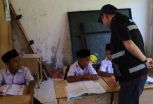 Seorang petugas instansi berbaju hitam dengan strip reflektif sedang berinteraksi dan berbicara dengan tiga siswa SD Aceh (mengenakan kemeja putih dan peci hitam) yang duduk di bangku sekolah kayu di sebuah ruang kelas yang menua dan berantakan. Siswa di tengah membuka buku pelajaran, sementara satu siswa di kiri tersenyum. Teks alt deskriptif: gambar interaksi petugas dan siswa di ruang kelas yang menua.