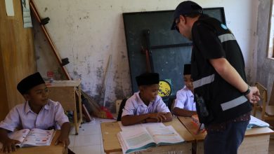 Seorang petugas instansi berbaju hitam dengan strip reflektif sedang berinteraksi dan berbicara dengan tiga siswa SD Aceh (mengenakan kemeja putih dan peci hitam) yang duduk di bangku sekolah kayu di sebuah ruang kelas yang menua dan berantakan. Siswa di tengah membuka buku pelajaran, sementara satu siswa di kiri tersenyum. Teks alt deskriptif: gambar interaksi petugas dan siswa di ruang kelas yang menua.