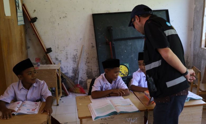 Seorang petugas instansi berbaju hitam dengan strip reflektif sedang berinteraksi dan berbicara dengan tiga siswa SD Aceh (mengenakan kemeja putih dan peci hitam) yang duduk di bangku sekolah kayu di sebuah ruang kelas yang menua dan berantakan. Siswa di tengah membuka buku pelajaran, sementara satu siswa di kiri tersenyum. Teks alt deskriptif: gambar interaksi petugas dan siswa di ruang kelas yang menua.