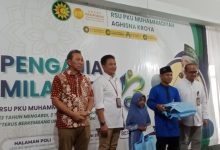 Sekelompok pria berfoto di atas panggung di depan spanduk acara di RSU PKU Muhammadiyah Aghisna Kroya, dengan salah satu pria memegang kotak berwarna biru yang mungkin berisi bingkisan.