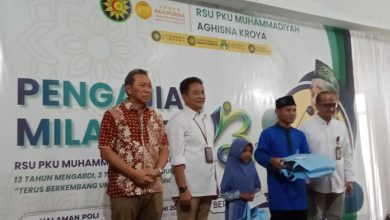 Sekelompok pria berfoto di atas panggung di depan spanduk acara di RSU PKU Muhammadiyah Aghisna Kroya, dengan salah satu pria memegang kotak berwarna biru yang mungkin berisi bingkisan.