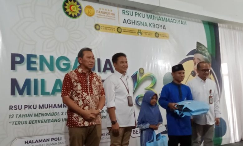 Sekelompok pria berfoto di atas panggung di depan spanduk acara di RSU PKU Muhammadiyah Aghisna Kroya, dengan salah satu pria memegang kotak berwarna biru yang mungkin berisi bingkisan.