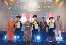 Sekelompok siswa dan guru berdiri di atas panggung dengan latar belakang layar LED besar. Layar tersebut menampilkan teks dalam bahasa Indonesia, termasuk "NILAI TERTINGGI TKA" dan "SMP MUHAMMADIYAH PROGRAM KHUSUS KOTTABARAT SURAKARTA." Di tengah, terdapat foto-foto siswa berprestasi. Dua siswa laki-laki dan satu siswa perempuan yang mengenakan topi toga dan jubah wisuda, masing-masing memegang piala dan piagam. Tiga orang dewasa berdiri di antara mereka; seorang wanita berhijab di sebelah kiri, seorang wanita di tengah, dan seorang pria di sebelah kanan. Dua pria tambahan berada di kedua ujung kelompok tersebut. Seluruh kelompok berpose menghadap kamera.
