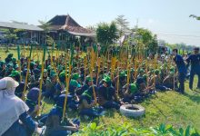 Ratusan siswa SD Muh PK Banyudono mengenakan seragam hijau Supercamp PPL-2 sedang duduk rapi dan memegang batang kayu untuk kegiatan penanaman pohon dalam program pendidikan karakter berbasis lingkungan di Refresh River Tubing Janti, Klaten. Dua mentor berbaju biru memberikan instruksi di depan.