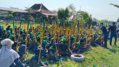 Ratusan siswa SD Muh PK Banyudono mengenakan seragam hijau Supercamp PPL-2 sedang duduk rapi dan memegang batang kayu untuk kegiatan penanaman pohon dalam program pendidikan karakter berbasis lingkungan di Refresh River Tubing Janti, Klaten. Dua mentor berbaju biru memberikan instruksi di depan.