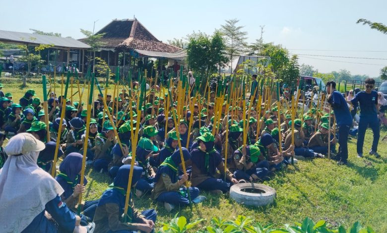 Ratusan siswa SD Muh PK Banyudono mengenakan seragam hijau Supercamp PPL-2 sedang duduk rapi dan memegang batang kayu untuk kegiatan penanaman pohon dalam program pendidikan karakter berbasis lingkungan di Refresh River Tubing Janti, Klaten. Dua mentor berbaju biru memberikan instruksi di depan.