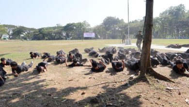 Puluhan mahasiswa PPG Unimus berpakaian hitam sedang melakukan latihan merayap di tanah berdebu di Lapangan Tembak Kodam IV/Diponegoro. Di latar belakang, tampak area lapangan rumput yang luas, tiang bendera, spanduk bertuliskan 'BELA NEGARA', serta instruktur TNI yang mengawasi jalannya latihan.