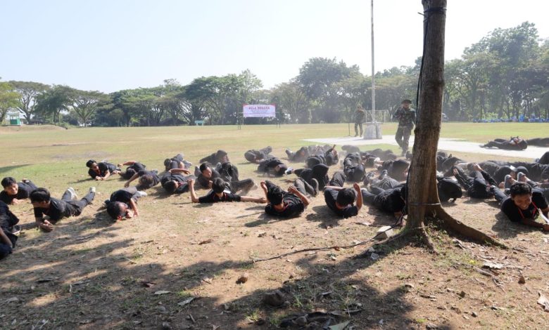 Puluhan mahasiswa PPG Unimus berpakaian hitam sedang melakukan latihan merayap di tanah berdebu di Lapangan Tembak Kodam IV/Diponegoro. Di latar belakang, tampak area lapangan rumput yang luas, tiang bendera, spanduk bertuliskan 'BELA NEGARA', serta instruktur TNI yang mengawasi jalannya latihan.