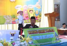 Seorang pria berpeci sedang mementaskan wayang golek pitutur di atas panggung dengan latar belakang spanduk anak-anak saleh yang berisi kutipan Arab.