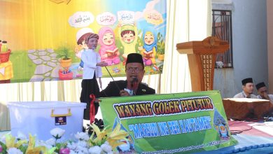 Seorang pria berpeci sedang mementaskan wayang golek pitutur di atas panggung dengan latar belakang spanduk anak-anak saleh yang berisi kutipan Arab.