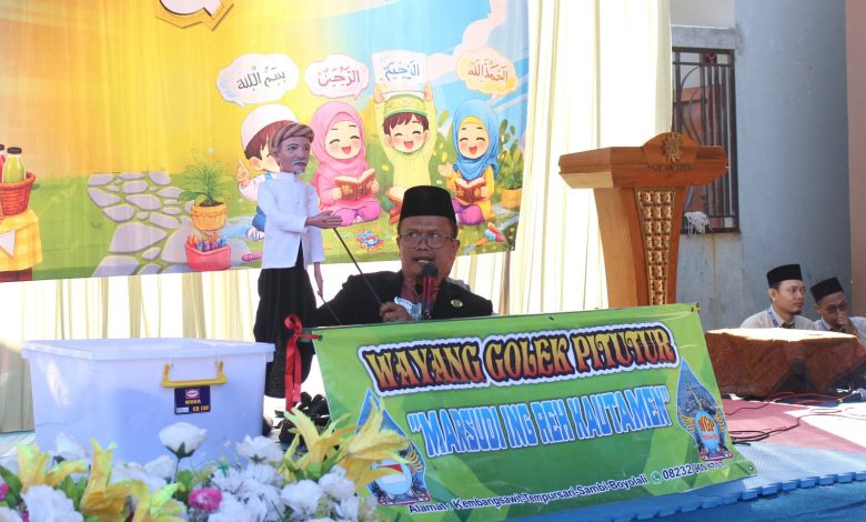Seorang pria berpeci sedang mementaskan wayang golek pitutur di atas panggung dengan latar belakang spanduk anak-anak saleh yang berisi kutipan Arab.