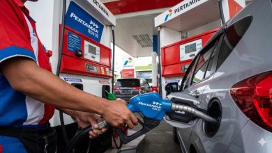 Petugas SPBU Pertamina berbaju merah kombinasi biru sedang memasukkan nosel biru Pertamax RON 92 ke dalam tangki bensin mobil pribadi berwarna perak.