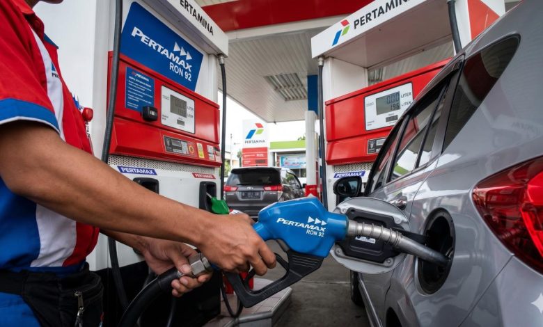 Petugas SPBU Pertamina berbaju merah kombinasi biru sedang memasukkan nosel biru Pertamax RON 92 ke dalam tangki bensin mobil pribadi berwarna perak.