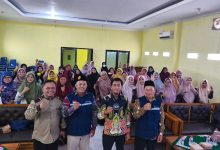 Empat orang pimpinan pria berdiri mengepalkan tangan di depan puluhan jemaah perempuan berhijab yang duduk rapi dalam sebuah ruang pertemuan atau aula berdidinding kuning.