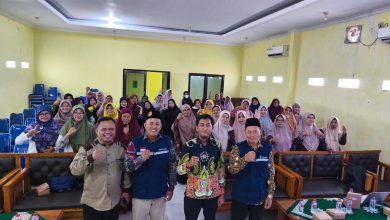 Empat orang pimpinan pria berdiri mengepalkan tangan di depan puluhan jemaah perempuan berhijab yang duduk rapi dalam sebuah ruang pertemuan atau aula berdidinding kuning.