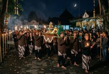 Prosesi kirab pusaka dan gunungan oleh masyarakat Jawa berpakaian adat beskap sambil membawa obor dan lilin pada Malam Satu Suro di bawah sinar bulan sabit.