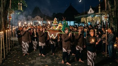 Prosesi kirab pusaka dan gunungan oleh masyarakat Jawa berpakaian adat beskap sambil membawa obor dan lilin pada Malam Satu Suro di bawah sinar bulan sabit.