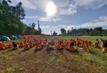 Puluhan siswa berseragam olahraga oranye duduk berkelompok di lapangan bumi perkemahan berlatar deretan tenda dome di bawah terik matahari.