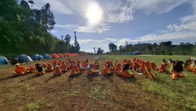 Puluhan siswa berseragam olahraga oranye duduk berkelompok di lapangan bumi perkemahan berlatar deretan tenda dome di bawah terik matahari.