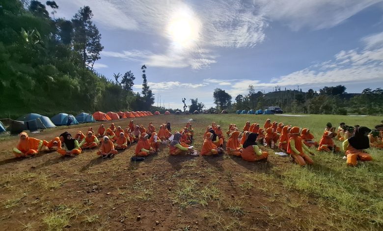 Puluhan siswa berseragam olahraga oranye duduk berkelompok di lapangan bumi perkemahan berlatar deretan tenda dome di bawah terik matahari.