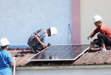 Tiga orang teknisi berhelm putih sedang memasang instalasi panel surya di atas atap seng gedung sekolah.