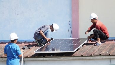 Tiga orang teknisi berhelm putih sedang memasang instalasi panel surya di atas atap seng gedung sekolah.