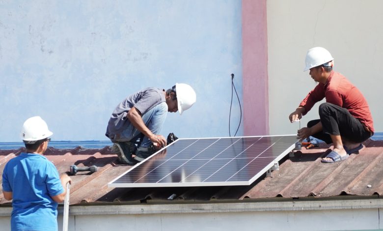 Tiga orang teknisi berhelm putih sedang memasang instalasi panel surya di atas atap seng gedung sekolah.