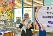 Seorang pembicara wanita berhijab sedang berbicara di depan peserta seminar parenting SD Muhammadiyah dengan spanduk acara di latar belakang.