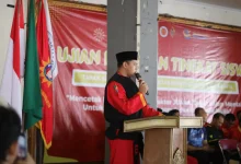 Sebuah gambar medium shot dari Dr. Eko Sudarmanto mengenakan seragam Tapak Suci berdiri di podium dan berbicara ke mikrofon.