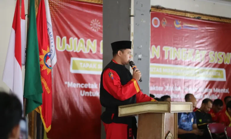 Sebuah gambar medium shot dari Dr. Eko Sudarmanto mengenakan seragam Tapak Suci berdiri di podium dan berbicara ke mikrofon.