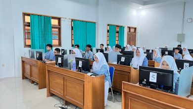Siswa-siswi SMA mengenakan seragam batik biru sedang fokus mengikuti ujian berbasis komputer atau ANBK di laboratorium komputer sekolah.