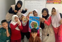 Mahasiswi KKN Internasional UMS bersama anak-anak perantauan di Malaysia memegang poster panduan porsi Isi Piringku Kemenkes di depan papan tulis 10 Pedoman Gizi Seimbang.